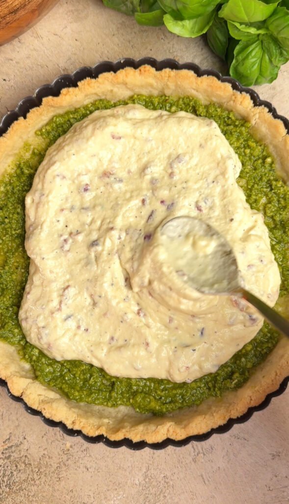 Ricotta, Parmesan, and bacon filling being spread over pesto layer inside golden tart crust