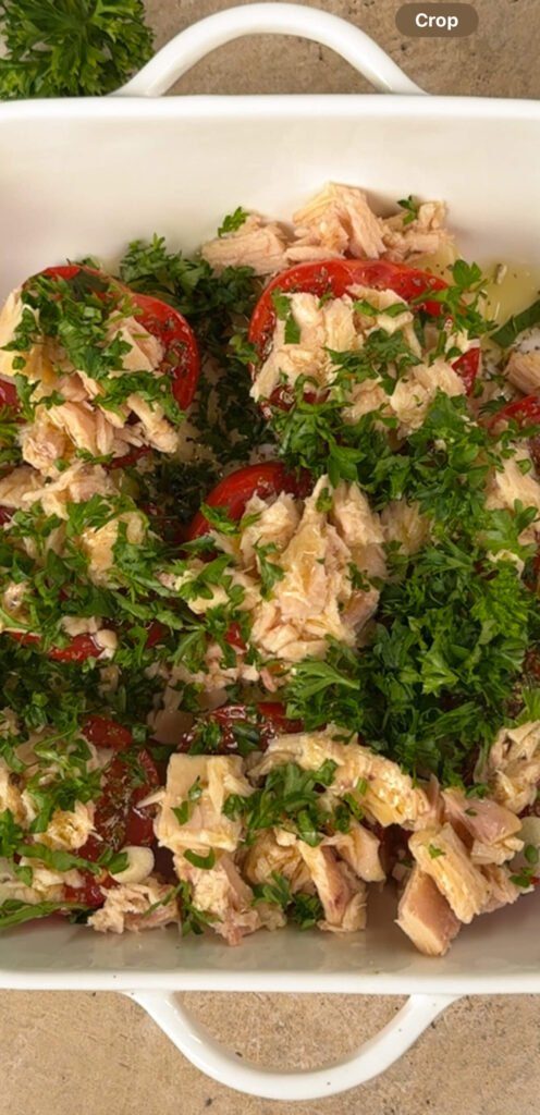 White baking dish filled with roasted Campari tomatoes, flaked tuna, chopped parsley, olive oil, and garlic — the savory topping for crostini before baking.