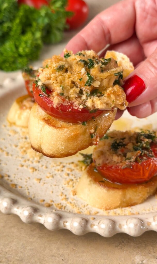 Hand holding a crostini topped with roasted tomato, tuna, herbs, and Parmesan crumbs — a golden, juicy bite of Mediterranean flavor.