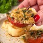 Hand holding a crostini topped with roasted tomato, tuna, herbs, and Parmesan crumbs — a golden, juicy bite of Mediterranean flavor.