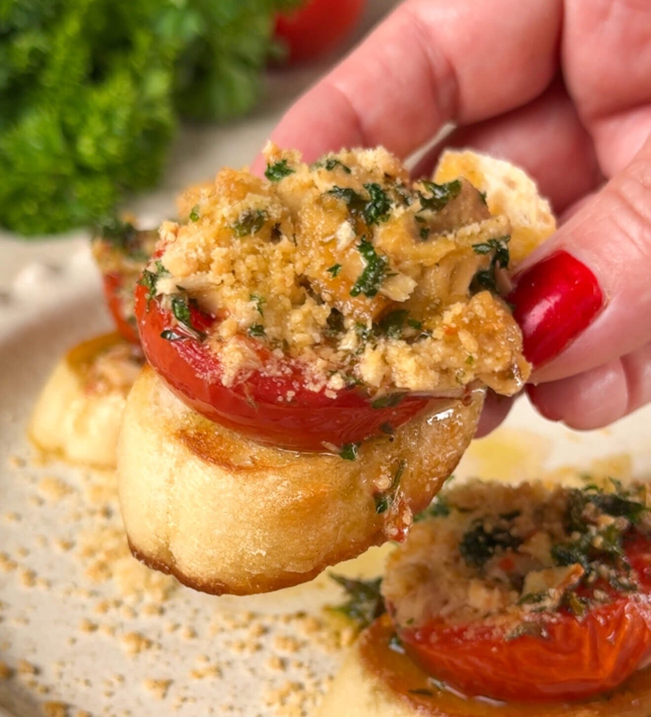 Hand holding a crostini topped with roasted tomato, tuna, herbs, and Parmesan crumbs — a golden, juicy bite of Mediterranean flavor.