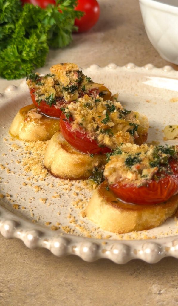 Three golden crostini topped with roasted tomatoes, flaked tuna, parsley, and Parmesan crumbs on a beige ceramic plate — a simple, elegant appetizer.