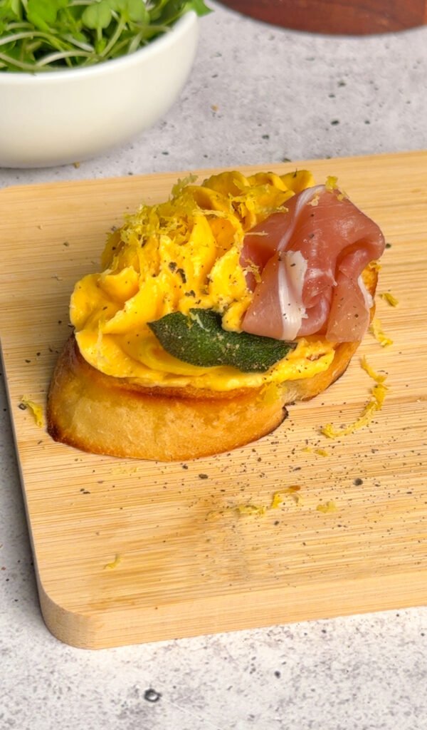 Butternut squash crostini topped with a prosciutto rose, crisp sage leaf, lemon zest, and black pepper on a wooden board.