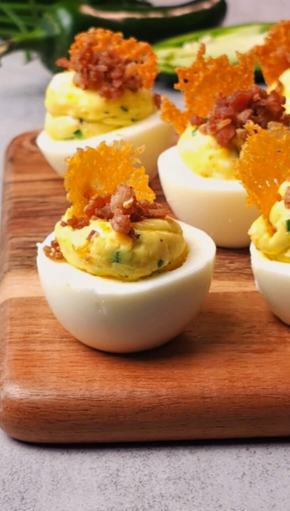 Plated jalapeño popper deviled eggs topped with crispy cheddar chips and crumbled bacon on a wooden serving board, with fresh jalapeños in the background