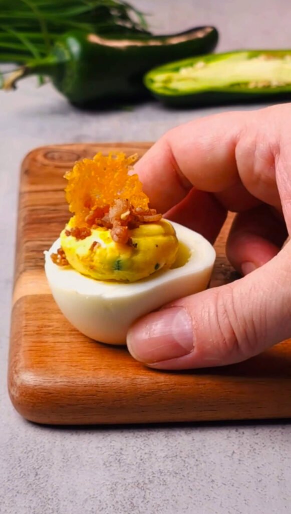 Hand placing a crispy cheddar chip on top of a jalapeño popper deviled egg filled with creamy yolk mixture and topped with bacon bits on a wooden board