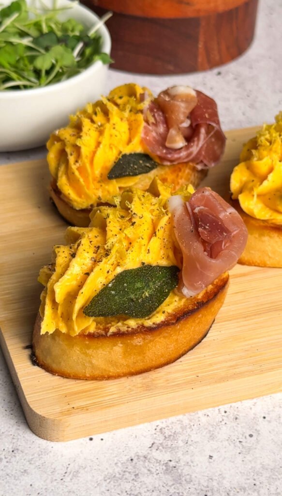 Close-up of butternut squash prosciutto crostini with piped mousse, fried sage, prosciutto roses, and lemon zest on toasted bread.