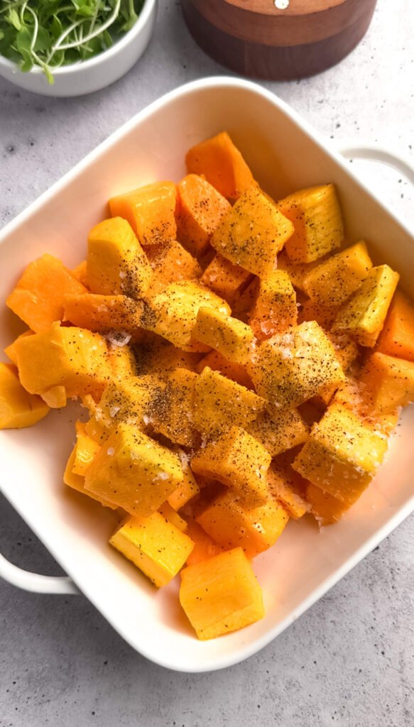 Cubed raw butternut squash seasoned with salt and black pepper in a white baking dish, ready to be roasted.