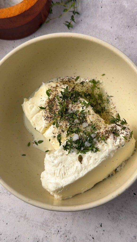 Soft goat cheese, cream cheese, fresh thyme, salt, and pepper in a bowl ready to be whipped into a creamy layer