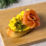 Close-up of one smoked salmon crostini with a salmon rose, golden mousse swirl, fresh dill, and crispy garlic-rubbed toast.
