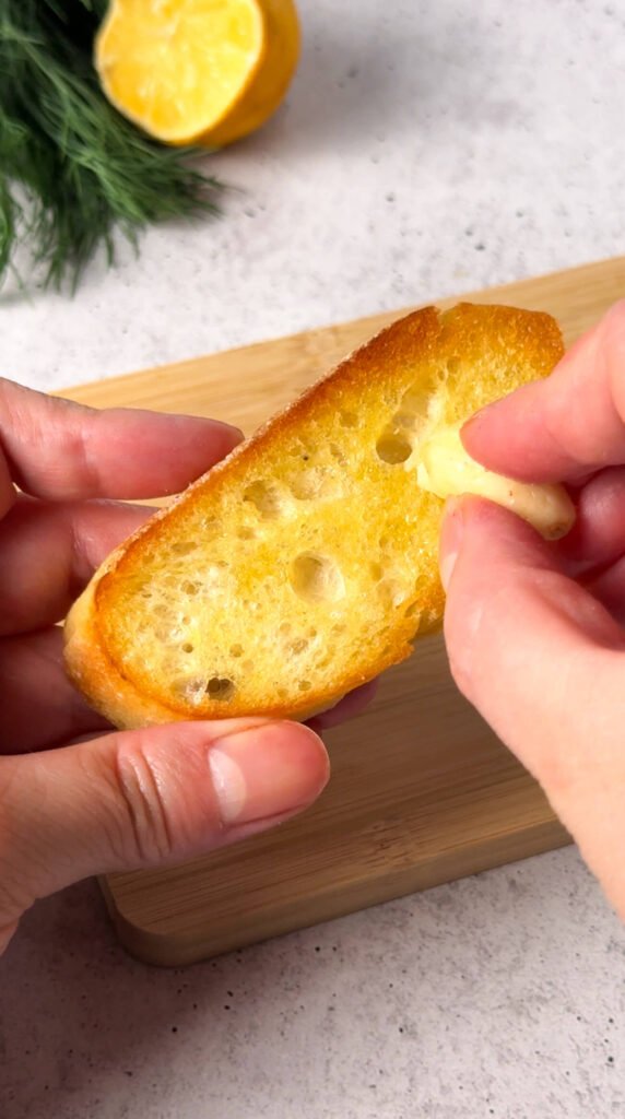 Hands rubbing a warm toasted baguette slice with a fresh garlic clove to infuse flavor before assembling smoked salmon crostini.
