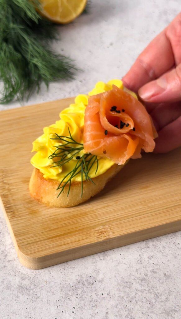 Close-up of one smoked salmon crostini with a salmon rose, golden mousse swirl, fresh dill, and crispy garlic-rubbed toast.