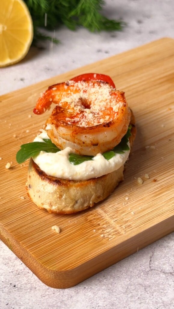 Finished shrimp crostini topped with seared shrimp, arugula, ricotta, cherry tomato, and grated Parmesan.