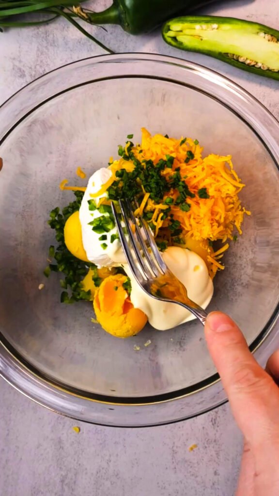 Mixing deviled egg filling with yolks, shredded cheddar, chopped chives, jalapeño, mayo, and cream cheese in a clear glass bowl — ingredients for jalapeño popper deviled eggs