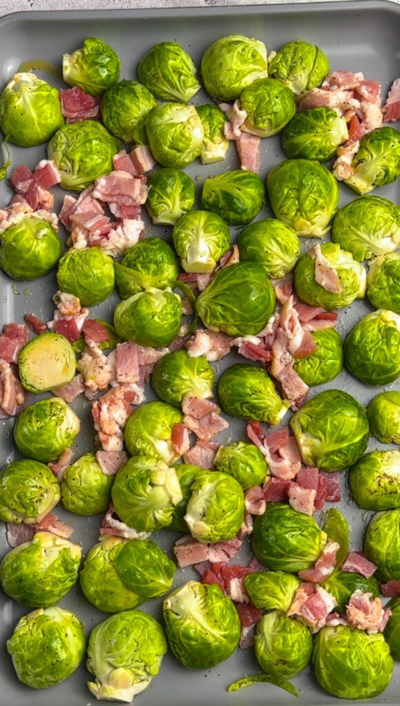 Raw Brussels sprouts and chopped bacon spread on a baking sheet before roasting at 400°F