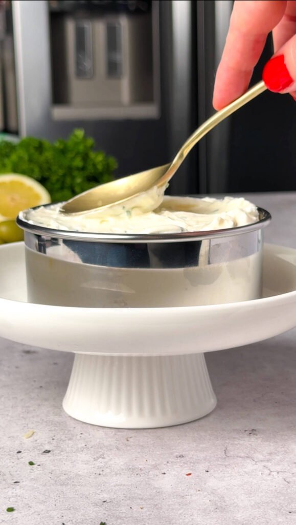 A hand using a spoon to spread whipped cream cheese into a metal ring mold, with fresh parsley and lemon visible in the background.