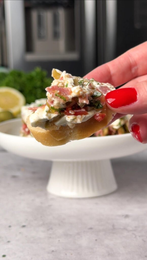 Close-up of a crostini topped with whipped cream cheese and a chopped salami and green olive mixture, held by a hand with red nail polish.