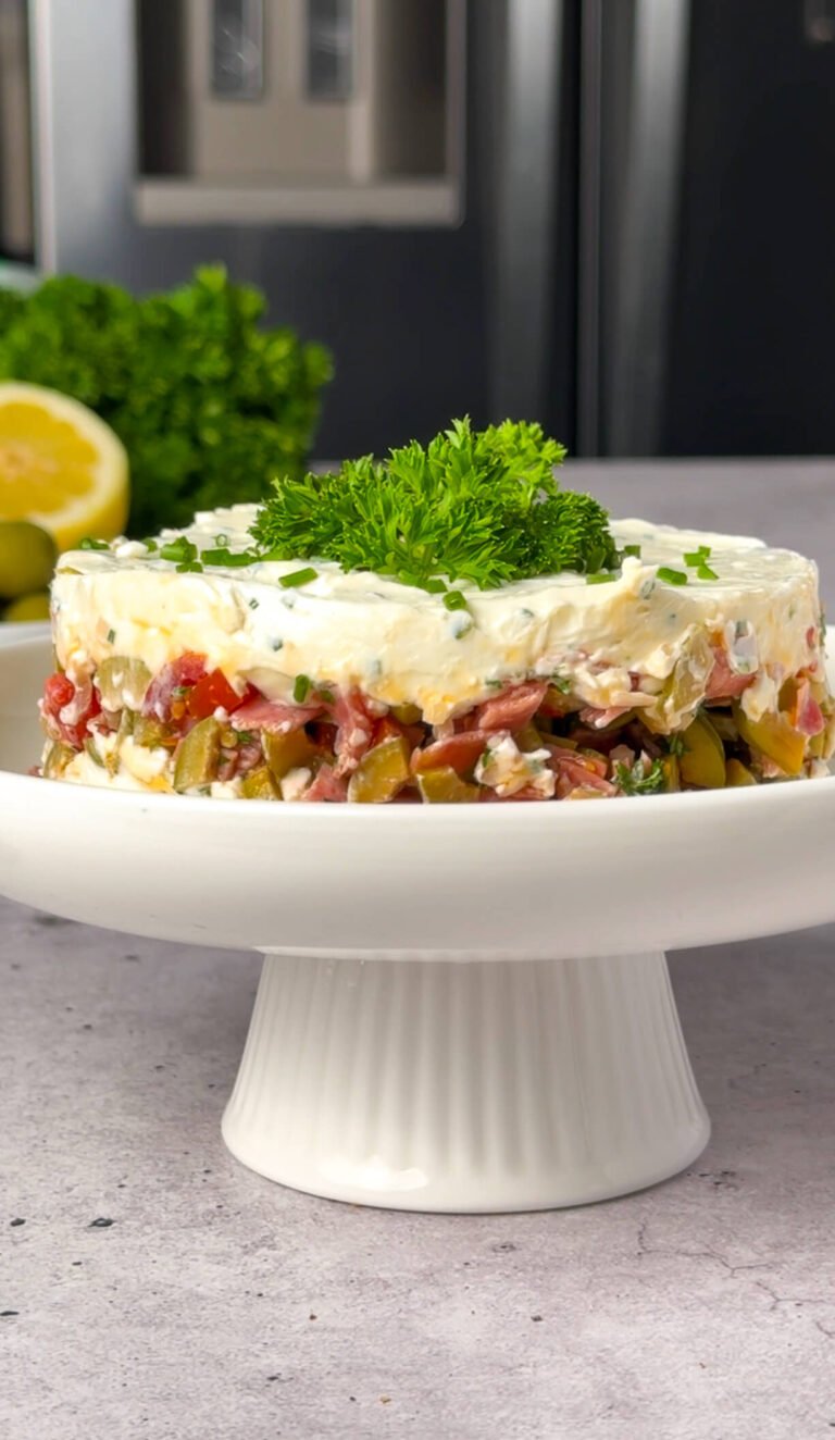 A layered salami and olive whipped cheese terrine on a white pedestal serving dish, topped with fresh parsley and chives.