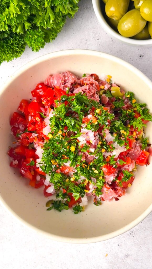 Finely chopped green olives, salami, cherry tomatoes, parsley, and shallot in a bowl, ready to be mixed.