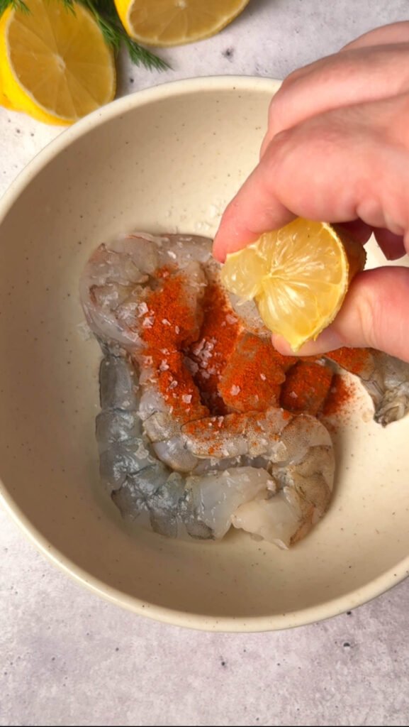 Raw shrimp in a bowl being seasoned with paprika, salt, and fresh lemon juice.