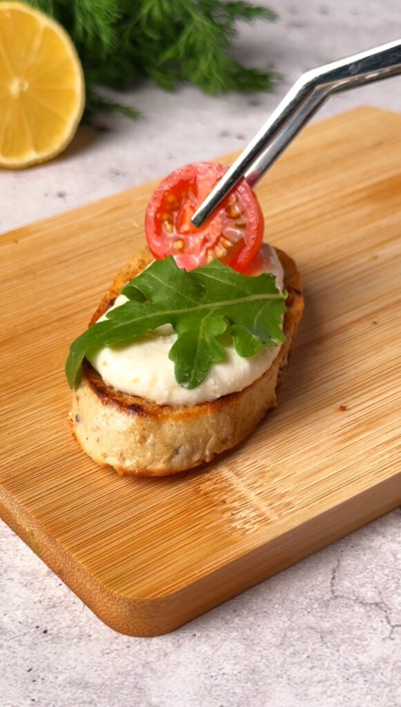 A cherry tomato slice being placed on top of arugula and ricotta on toasted bread.