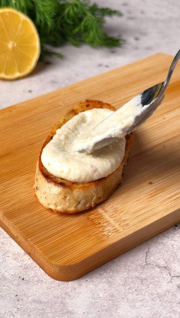 Whipped ricotta being spread onto a toasted baguette slice on a wooden board.