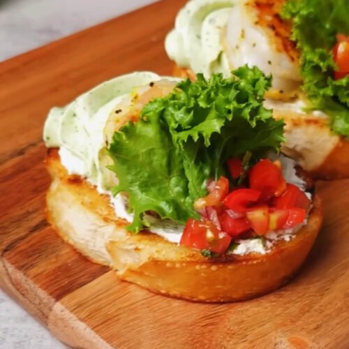 A finished shrimp crostini on a wooden board topped with basil whipped cream cheese, a sautéed shrimp, a frilly green lettuce leaf, and a spoonful of diced cherry tomatoes.