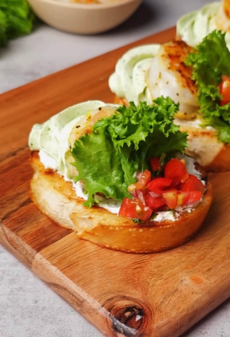 A finished shrimp crostini on a wooden board topped with basil whipped cream cheese, a sautéed shrimp, a frilly green lettuce leaf, and a spoonful of diced cherry tomatoes.