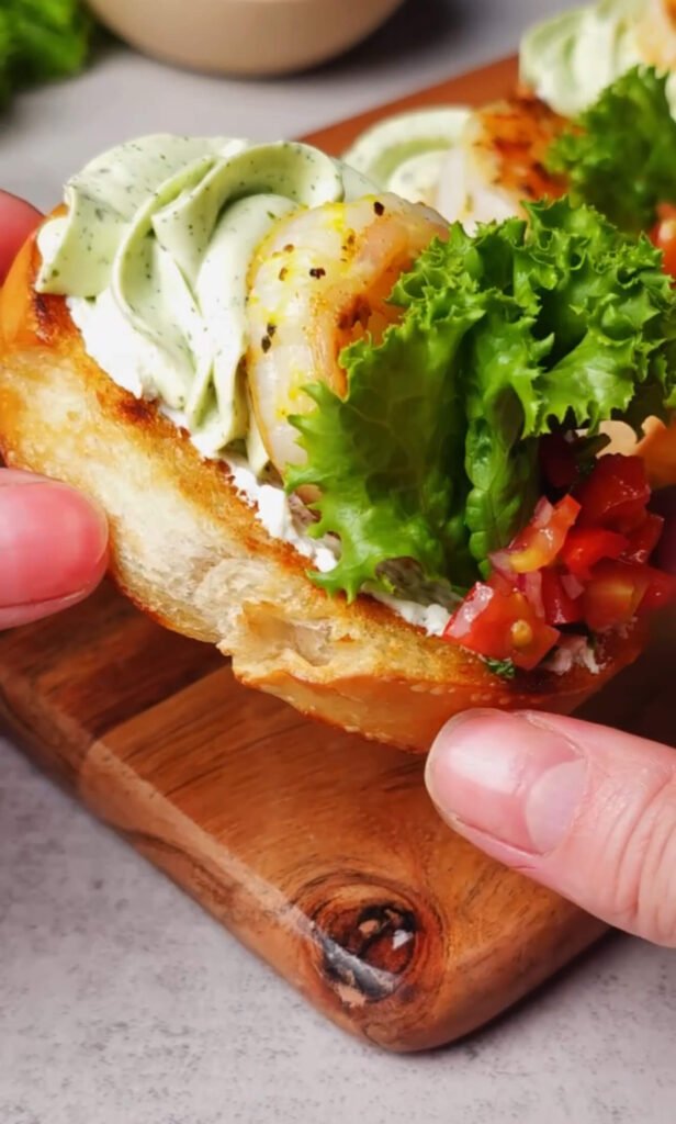 A hand holds a fully assembled shrimp crostini with a basil cream cheese swirl, golden shrimp, fresh lettuce, and tomato–shallot topping on a toasted baguette slice.