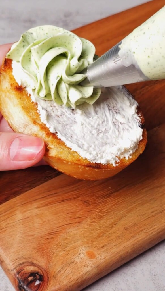 A hand holds a toasted baguette slice spread with plain cream cheese while a piping bag swirls a decorative rosette of pale green basil whipped cream cheese on top.