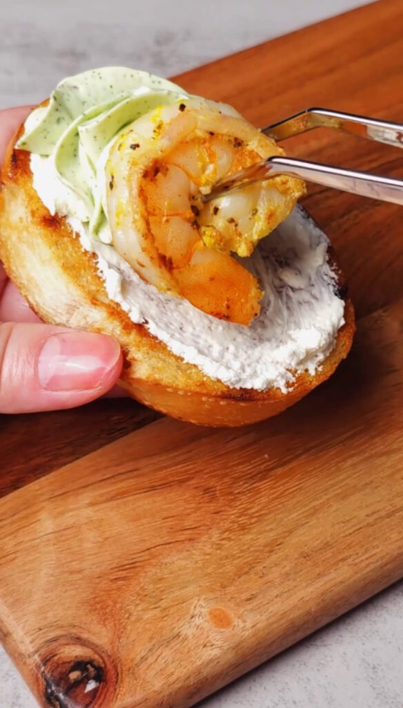 A cooked shrimp is being placed onto a toasted crostini topped with a swirl of basil whipped cream cheese and a thin layer of plain cream cheese.