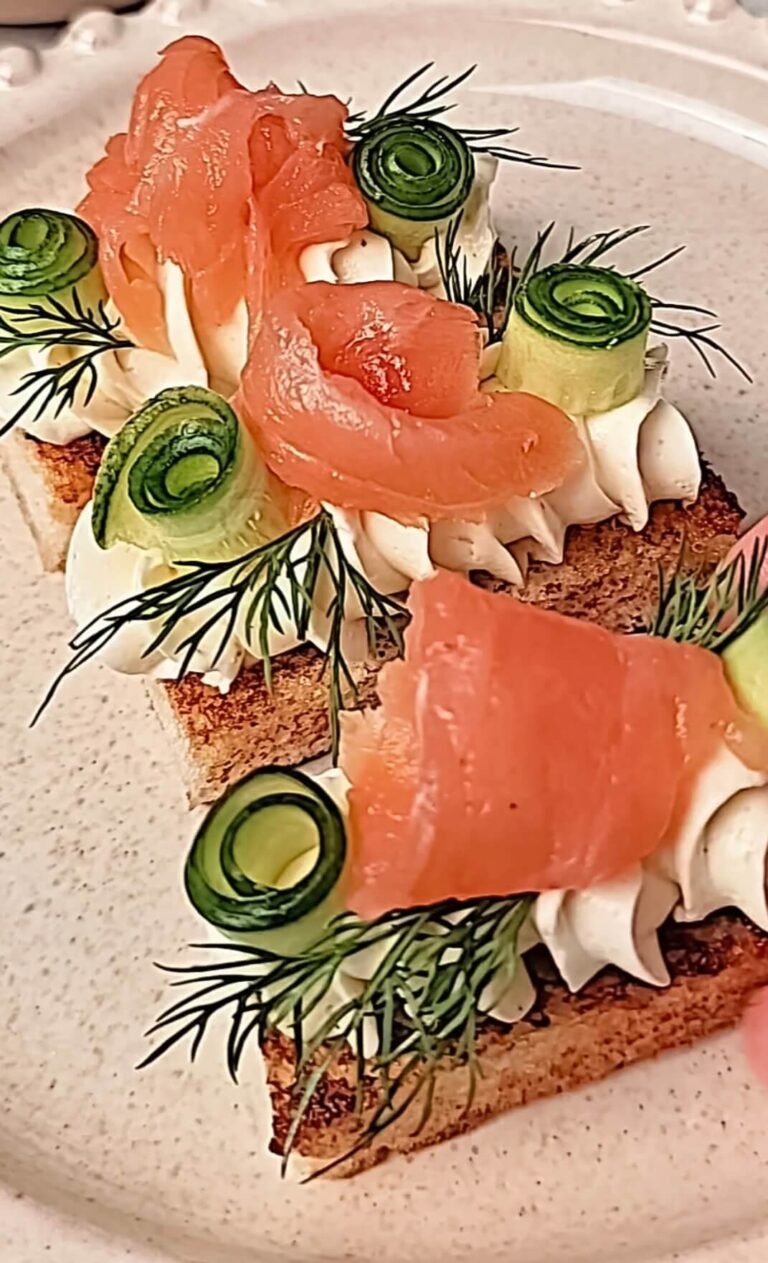 Several smoked salmon crostini arranged on a plate, each topped with whipped cream cheese, cucumber ribbon curls, and fresh dill for serving.