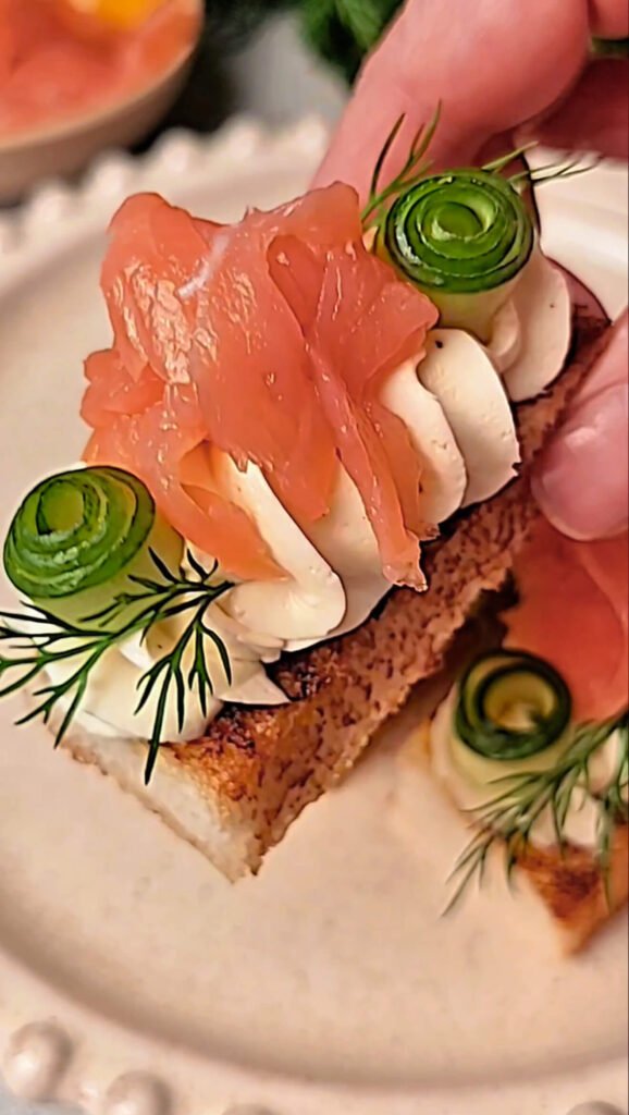 Hand picking up a smoked salmon and whipped cream cheese crostini garnished with cucumber ribbons and dill, showing the bite-size scale and texture details.