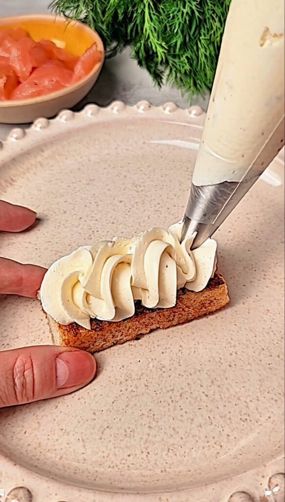 Whipped cream cheese being piped in soft decorative swirls onto a small toasted rectangle of bread, with fresh dill and smoked salmon visible in the background.