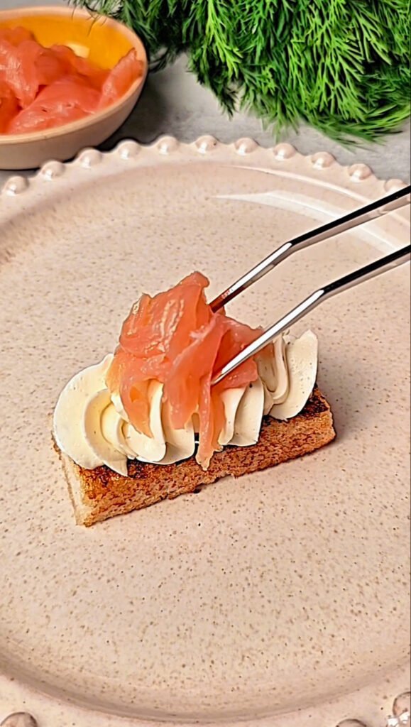 Smoked salmon being carefully placed onto piped cream cheese on toast using kitchen tweezers, highlighting the precise presentation.