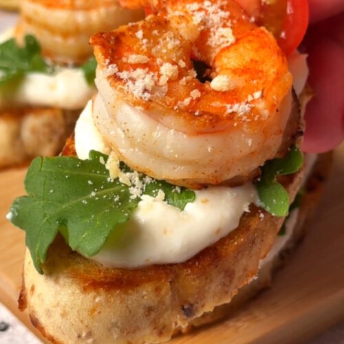 Hand picking up a Zesty Shrimp Crostini topped with whipped ricotta, arugula, cherry tomato, and seared shrimp.