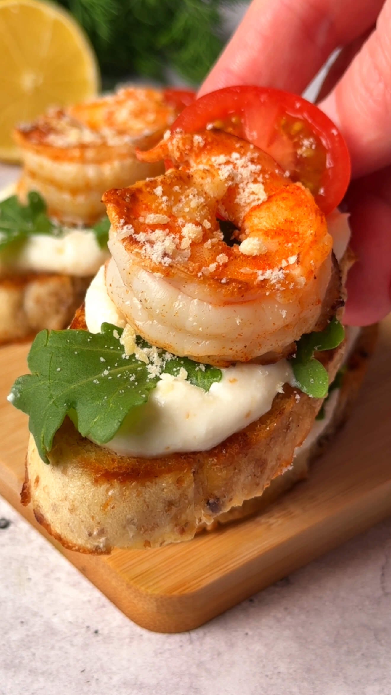 Hand picking up a Zesty Shrimp Crostini topped with whipped ricotta, arugula, cherry tomato, and seared shrimp.