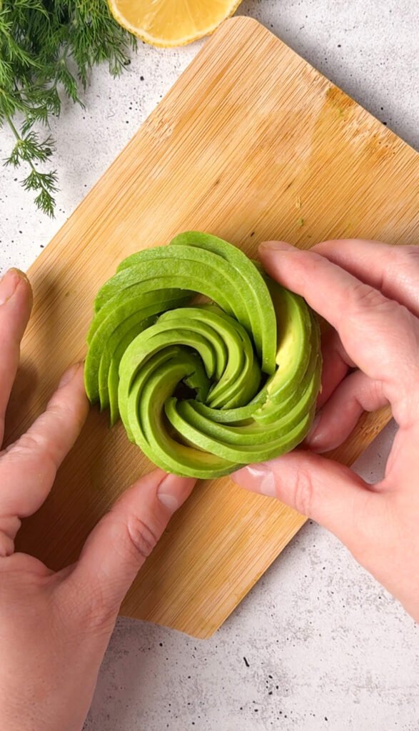 Hands shaping thin avocado slices into a tight avocado rose on a wooden board
