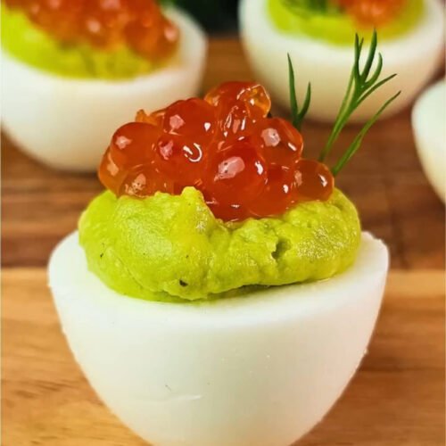 Close-up of an avocado deviled egg topped with red caviar and fresh dill on a wooden board