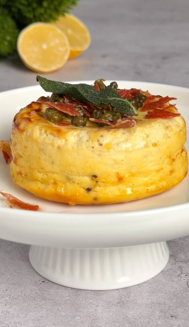 Baked ricotta appetizer unmolded and topped with crispy prosciutto, fried sage, and capers, served warm on a white pedestal plate