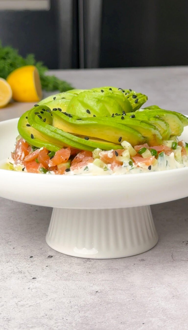Smoked salmon tartare stack topped with an avocado rose and sprinkled with nigella seeds, served on a white pedestal dish