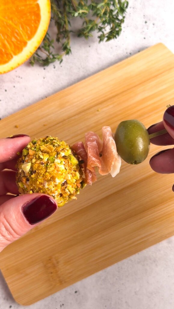 Assembled goat cheese prosciutto bonbon with pistachio crust, folded prosciutto, and green olive on a cocktail pick