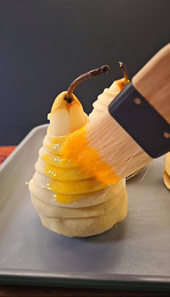 A peeled Bosc pear wrapped in thin puff pastry strips being brushed with glossy egg wash, ready to bake on a sheet pan.