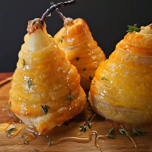Golden puff pastry–wrapped pears drizzled with honey and garnished with fresh thyme, displayed on a wooden board