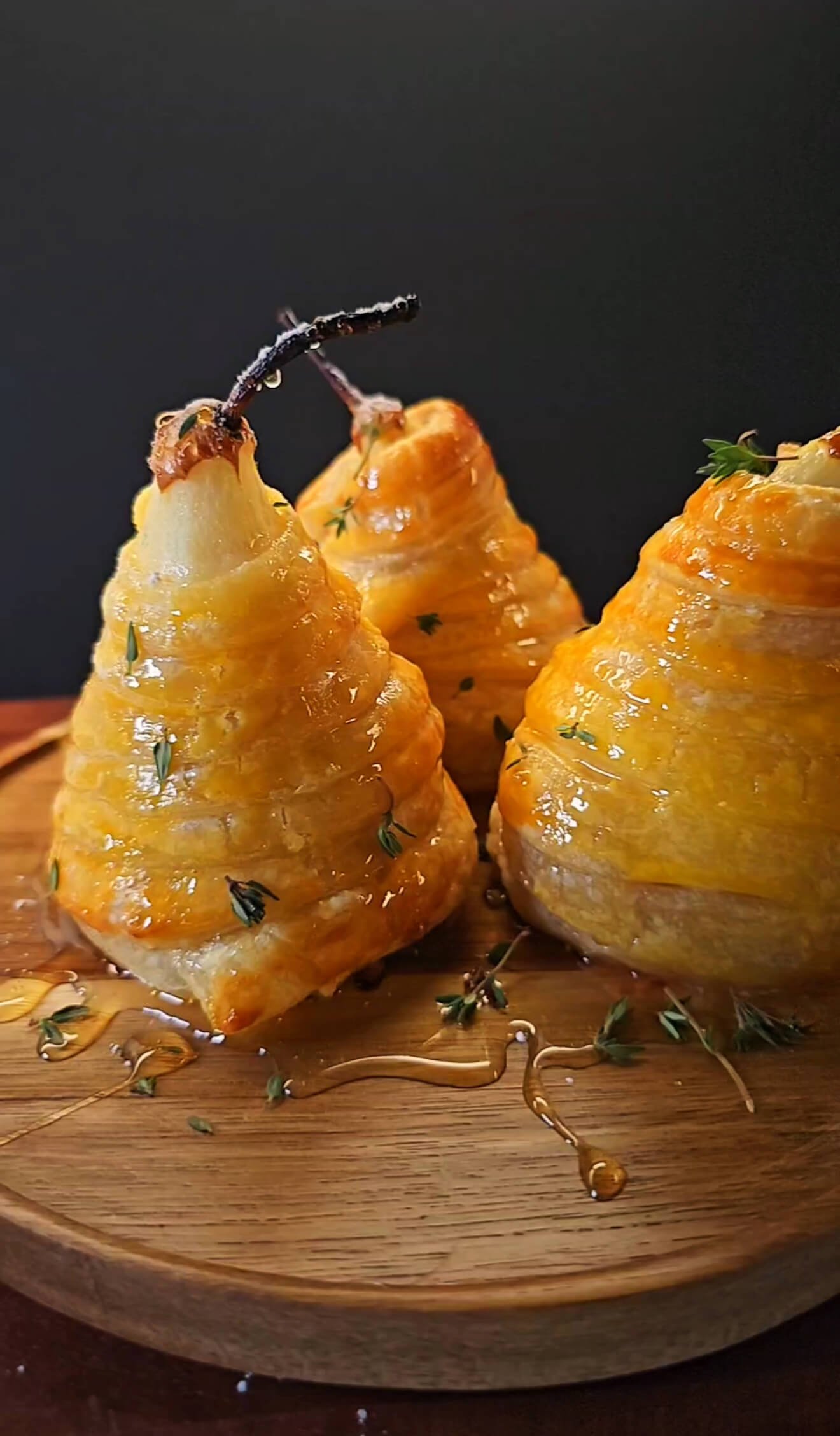 Golden puff pastry–wrapped pears drizzled with honey and garnished with fresh thyme, displayed on a wooden board