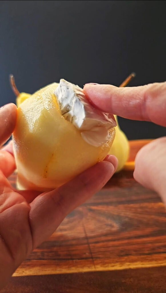 Close-up of a peeled pear with a cube of creamy Brie cheese being tucked into the hollowed center before wrapping.