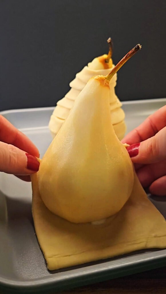A peeled pear placed upright on a square of puff pastry as hands gently wrap the dough around the fruit before spiraling.