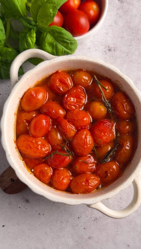 A bowl of freshly roasted cherry tomatoes, soft, glossy, and caramelized, with rosemary sprigs visible in the warm tomato oil.