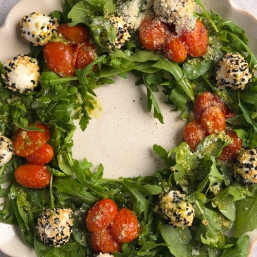 A fully assembled Sesame Mozzarella Balls Wreath on a scalloped platter, topped with roasted cherry tomatoes, sesame-coated mozzarella balls, arugula, and Parmesan.