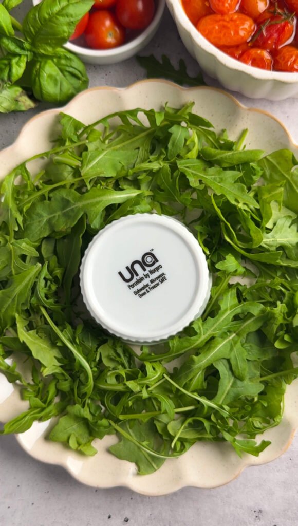 A round platter lined with fresh arugula arranged in a wreath shape, with a small bowl in the center to create the circle.