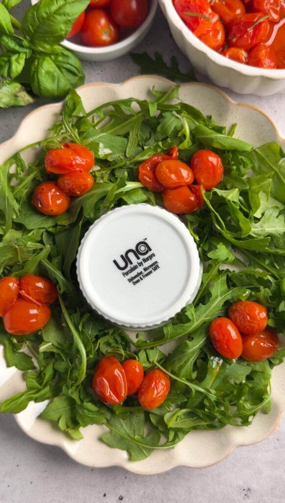 Arugula wreath base with clusters of glossy slow-roasted cherry tomatoes placed around the greens before final assembly.